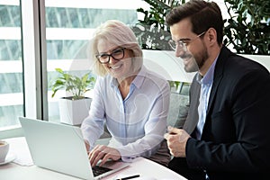 Happy middle aged leader working on computer with young colleague.