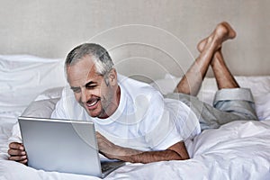 Happy middle aged man using laptop while lying in bed at home