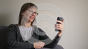 Happy middle aged mature woman holding smartphone having video chat, using social media apps in telephone sit on couch