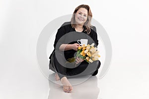 Happy middle-aged plump woman in black cardigan and leggings sitting, holding bouquet of yellow tulips and white cup.