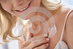 Happy middle aged woman applying cream moisturizing lotion on body skin.
