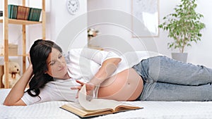 Happy middle-aged woman is eight months pregnant relaxing in bedroom, lying on bed and reading book