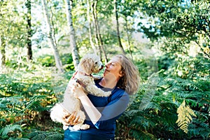 Happy middle-aged woman embracing her cockapoo puppy pet dog during walk outdoors in summer forest or park on sunset. Cute little