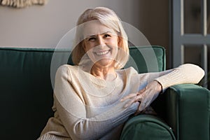 Happy middle aged woman looking at camera sit on sofa