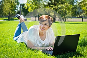 Happy middle aged woman lying on green grass using laptop computer in the park. adult man running outdoors. business