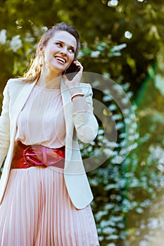 happy middle aged woman in pink dress and white jacket in the city talking on a smartphone