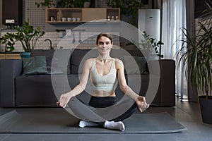 Happy middle-aged woman sitting in lotus position meditating, doing fitness in the morning at home, looking at camera