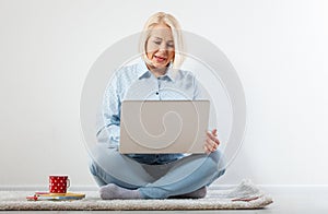 Happy middle aged woman sitting relaxed on the floor using laptop for entertainment. The concept of leisure and work with a cup of