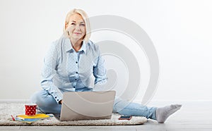 Happy middle aged woman sitting relaxed on the floor using laptop for entertainment. The concept of leisure and work with a cup of