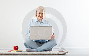 Happy middle aged woman sitting relaxed on the floor using laptop for entertainment. The concept of leisure and work with a cup of