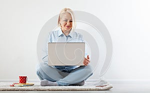 Happy middle aged woman sitting relaxed on the floor using laptop for entertainment. The concept of leisure and work with a cup of
