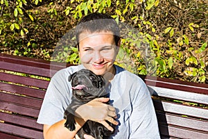 Happy middle-aged woman smiling enjoying her black pug dog on bench on park natural background