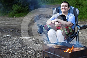 Happy middle-aged woman smiling, hagging black pug dog on tourist chair before fire in evening