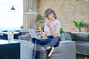 Happy middle aged woman using laptop, sitting on sofa at home