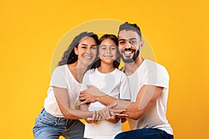 Happy Middle-Eastern Parents Hugging Their Kid Daughter On Yellow Background