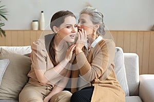 Happy middle mother whispering secret to her smiling daughter at home, gossiping, sharing secret