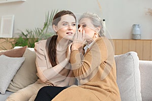 Happy middle mother whispering secret to her smiling daughter at home, gossiping, sharing secret