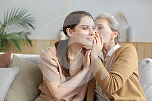 Happy middle mother whispering secret to her smiling daughter at home, gossiping, sharing secret.