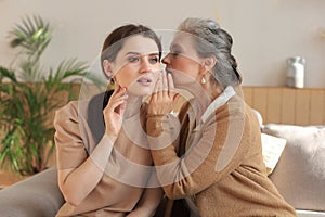 Happy middle mother whispering secret to her smiling daughter at home, gossiping, sharing secret.