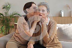 Happy middle mother whispering secret to her smiling daughter at home, gossiping, sharing secret.