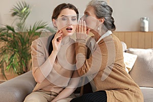 Happy middle mother whispering secret to her smiling daughter at home, gossiping, sharing secret.