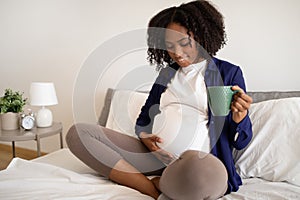 Happy millennial african american pregnant lady with big belly drinks cup of tea, feels baby move on bed