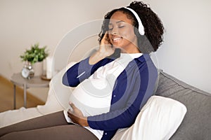 Happy millennial african american woman with big belly in headphones enjoys music in room interior