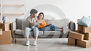 Happy millennial Asian couple with laptop sitting on couch among cardboard boxes and planning design of their new home