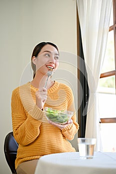 Happy millennial Asian woman having lunch, enjoys eating healthy salad vegetables mix at home