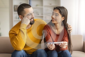 Happy millennial couple holding positive pregnancy test and embracing at home