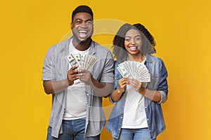 Happy millennial couple posing with lot of money, holding with dollar fans, orange panorama background with empty space