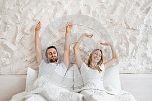 Happy millennial couple stretching on bed after waking up, feeling sleepy at home, copy space