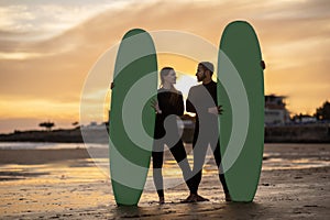 Happy millennial couple with surfboards standing on the beach on sunset time