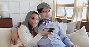 Happy millennial couple watching tv holding remote control on sofa