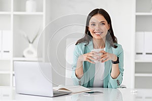 Happy millennial european businesswoman uses computer, drink coffee, enjoy work, relax in white office