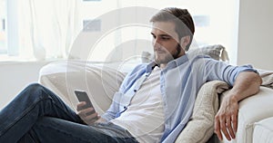 Happy millennial guy relaxing on sofa holding using smart phone