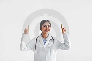 Happy millennial indian lady doctor in coat with phonendoscope raises hands and shows fingers up