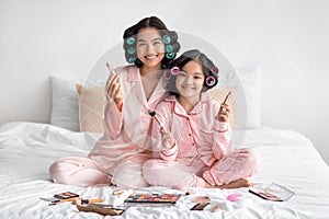Happy millennial japanese female and adolescent girl with curlers hold brushes sit on bed with cosmetics