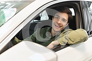Happy millennial man sitting on driver& x27;s seat of new automobile after purchase, looking out window and smiling at
