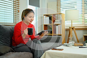 Happy millennial woman drinking hot tea on couch and surfing internet on digital tablet