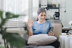 Happy millennial woman having video chat with friends using smart phone while sitting on sofa in living room at home.