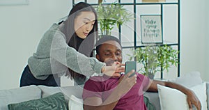 Happy mixed race couple talk laugh looking at smartphone using funny apps sit on couch. Smiling man and woman relaxing