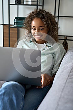 Happy mixed race female student sitting on soft couch at home typing on laptop.