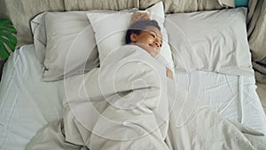 Happy mixed race girl is lying in bed and smiling then stretching arms, closing eyes and falling asleep. Resting people