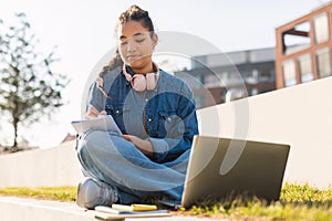 Happy mixed race teen lady writing something in notebook, sitting with laptop computer in park, studying outdoors