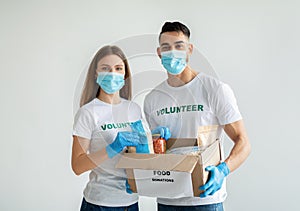 Happy mixed race volunteers in medical masks packing food donations in box standing over light wall and smiling