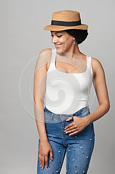 Happy mixed race woman in jeans and white tank top wearing canotier straw hat with hands in pockets