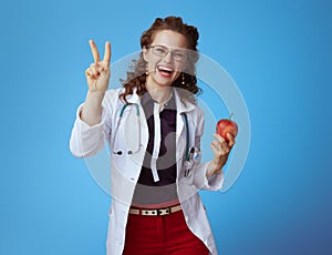 Happy modern doctor woman with apple showing victory on blue