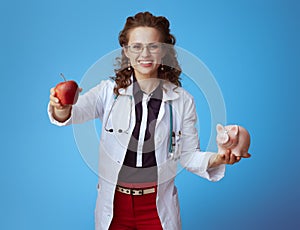 Happy modern doctor woman showing apple and piggy bank on blue
