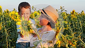 Happy mom and baby are looking at a bright yellow sunflower flower in the field at sunset. A little boy sitting on his
